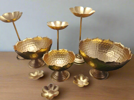 A set of gold-colored iron urli bowls with stands, displayed on a grass-like surface, against a blue background.
