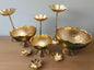 A set of gold-colored iron urli bowls with stands, displayed on a grass-like surface, against a blue background.